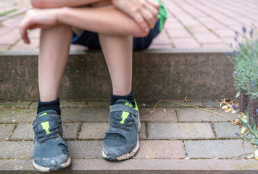 Ein Kind das am Gehsteig sitzt und man sieht nur die Füße mit kaputten Schuhen und die Arme die auf den Knien liegen