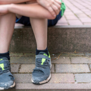 Ein Kind das am Gehsteig sitzt und man sieht nur die Füße mit kaputten Schuhen und die Arme die auf den Knien liegen